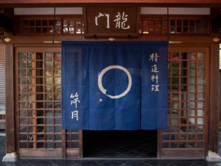 The restaurant entrance lies behind a blue noren curtain nestled behind some trees