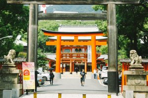 Pintu masuk kuil Ikuta dari jalan raya. Terlihat sebuah pintu/torii dari kayu, melewati jalan masuk/sando Anda akan bertemu lagi dengan torii berwarna merah.