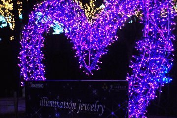 ตัวการ์ตูนน่ารักยืนต้อนรับคุณอยู่ตรงหน้างานประดับไฟรูปหัวใจที่มีชื่อว่า  'Illumination Jewelry'