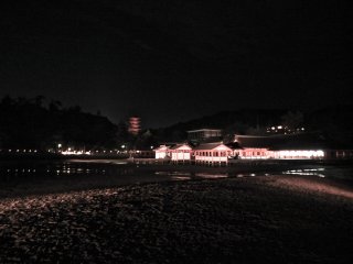 Vista de longe para o Santu&aacute;rio Itsukushima, flutuando na escurid&atilde;o, com o Templo Daisho-in por tr&aacute;s