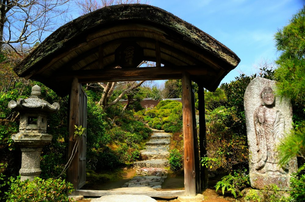 An arched roof of Hiwada-buki (thatched-roof using Japanese cypress bark)! An elaborate gate with a stone lantern and stone Buddha statue on each side.