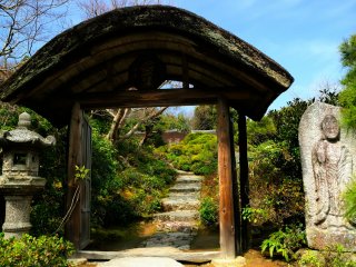 An arched roof of Hiwada-buki (thatched-roof using Japanese cypress bark)! An elaborate gate with a stone lantern and stone Buddha statue on each side.