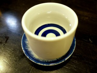 สาเกในถ้วยสีฟ้าขาวที่มีจานรอง ซึ่งถือว่าเป็นความคิดที่ดี ถ้าคุณทำตามธรรมเนียมญี่ปุ่นที่รินสาเกจนเต็มถึงปากถ้วย