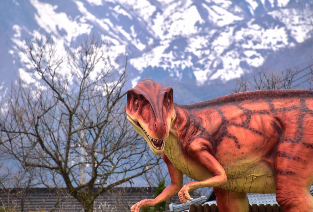 A dinosaur replica standing at the foot of Katsuyama Bridge with a snowy mountain in the backdrop