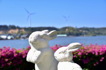 <p>Statues of rabbits in the garden of Kitagata Lakeside Hotel with pink azaleas, the lake and windmills in the background</p>