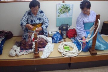 Ladies demonstrate different shibori techniques.  They are both wearing clothes in the pattern they are creating.