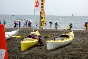 Kayaks lie in the sand, while in the background, the crowd survey weather conditions before the relay race begins