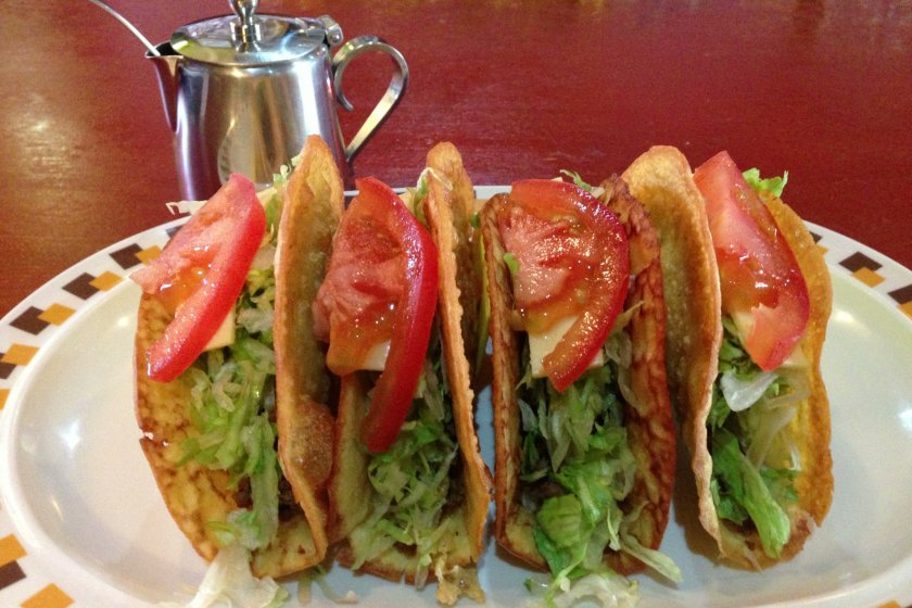 Okinawan tacos are prepared with rice-based taco shells, flavorable chili spiced beef, cheese, shreeded lettuce and tomatoes, and are made exactly the same each time; taco sauce is served in a coffee creamer pitcher with a spoon