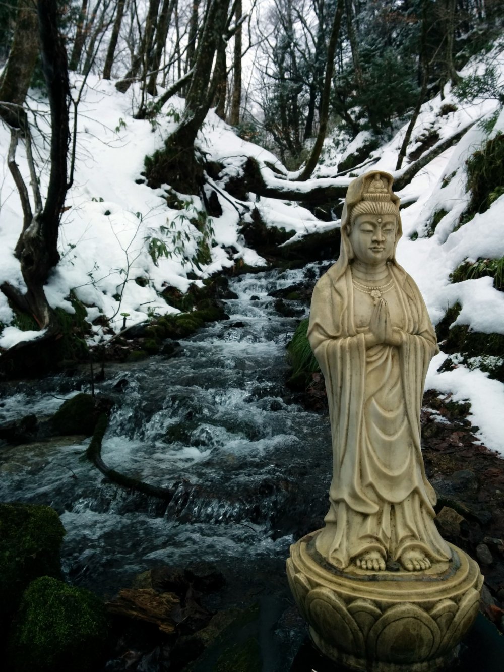 A Buddhist statue stands silently along an icy stream.